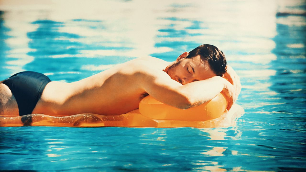 A man relaxing on an orange float in a clear blue swimming pool, wearing dark swim trunks.