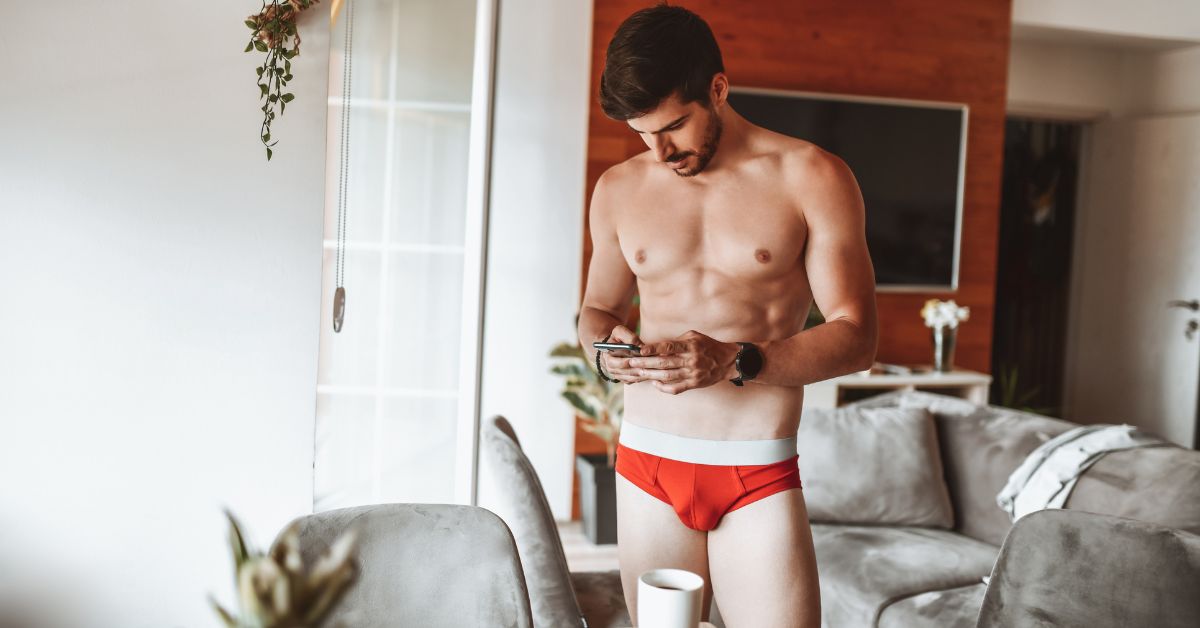 Man in red men's briefs using a smartphone in a modern living room.