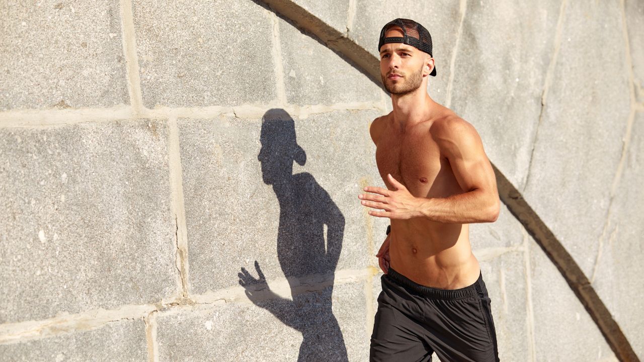 A fit man jogging shirtless outdoors, wearing black shorts and a backward cap.