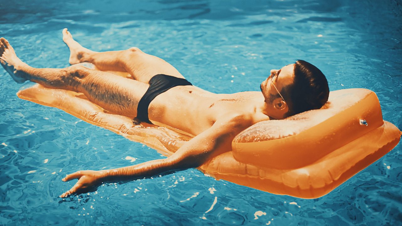 A man wearing black swim briefs relaxes on an orange pool float in a bright blue pool.