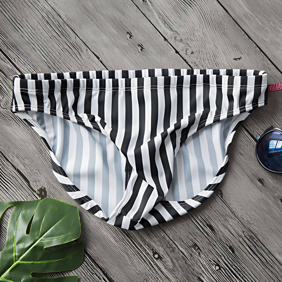 Flat lay of Black & White Vertical Stripe Swim Briefs on a wooden surface with tropical leaf and sunglasses.