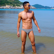 Man wearing Black & White Vertical Stripe Swim Briefs standing in shallow water with a scenic beach background.