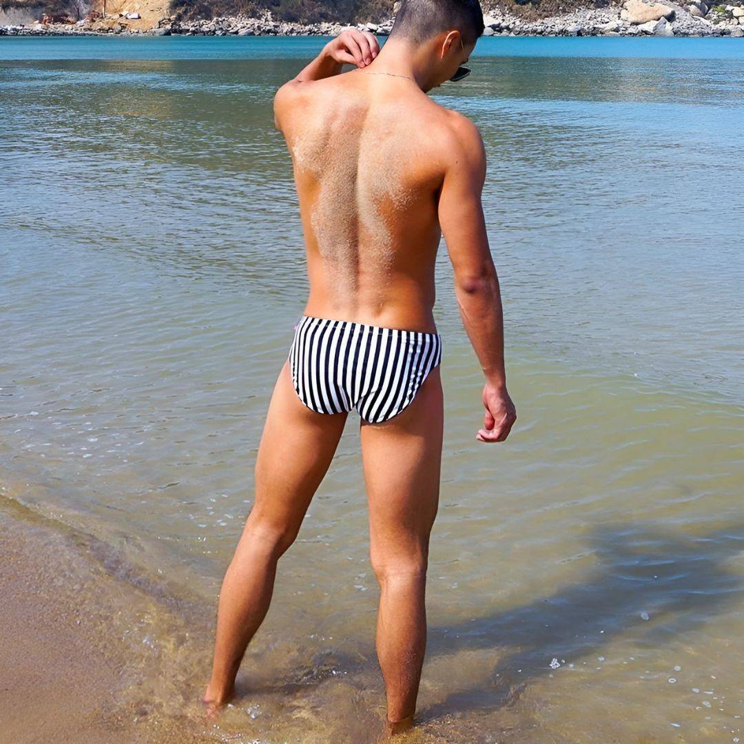 Rear view of Black & White Vertical Stripe Swim Briefs worn by a man at the beach, showcasing the swimwear's fit.