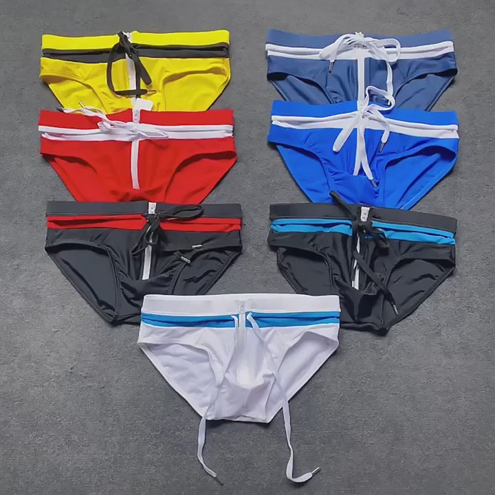 Collection of zipper front swim briefs in various colors, including yellow, blue, red, black, and white, arranged on a gray surface.