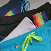 Variety of swim briefs in blue, black, white, and navy, each with a rainbow back pocket and adjustable drawstrings.